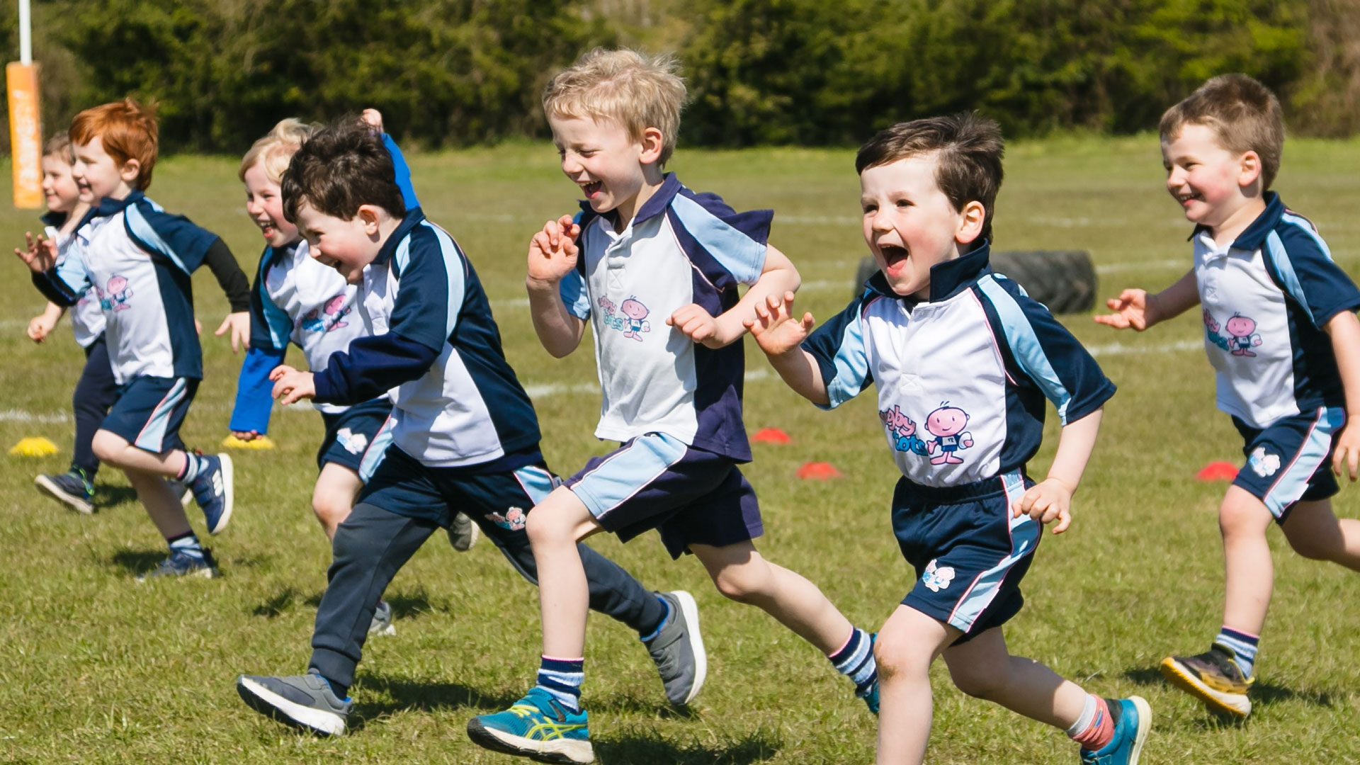 Children with happy faces running across a sports field during a Rugbytots session.